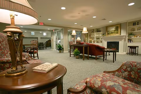 A spacious and well-lit living room area in a senior living facility featuring floral upholstered chairs, a wooden side table with a lamp and a book, a red sofa, a fireplace with a painting above it, built-in shelves with decorative items, and additional seating and tables in the background.
