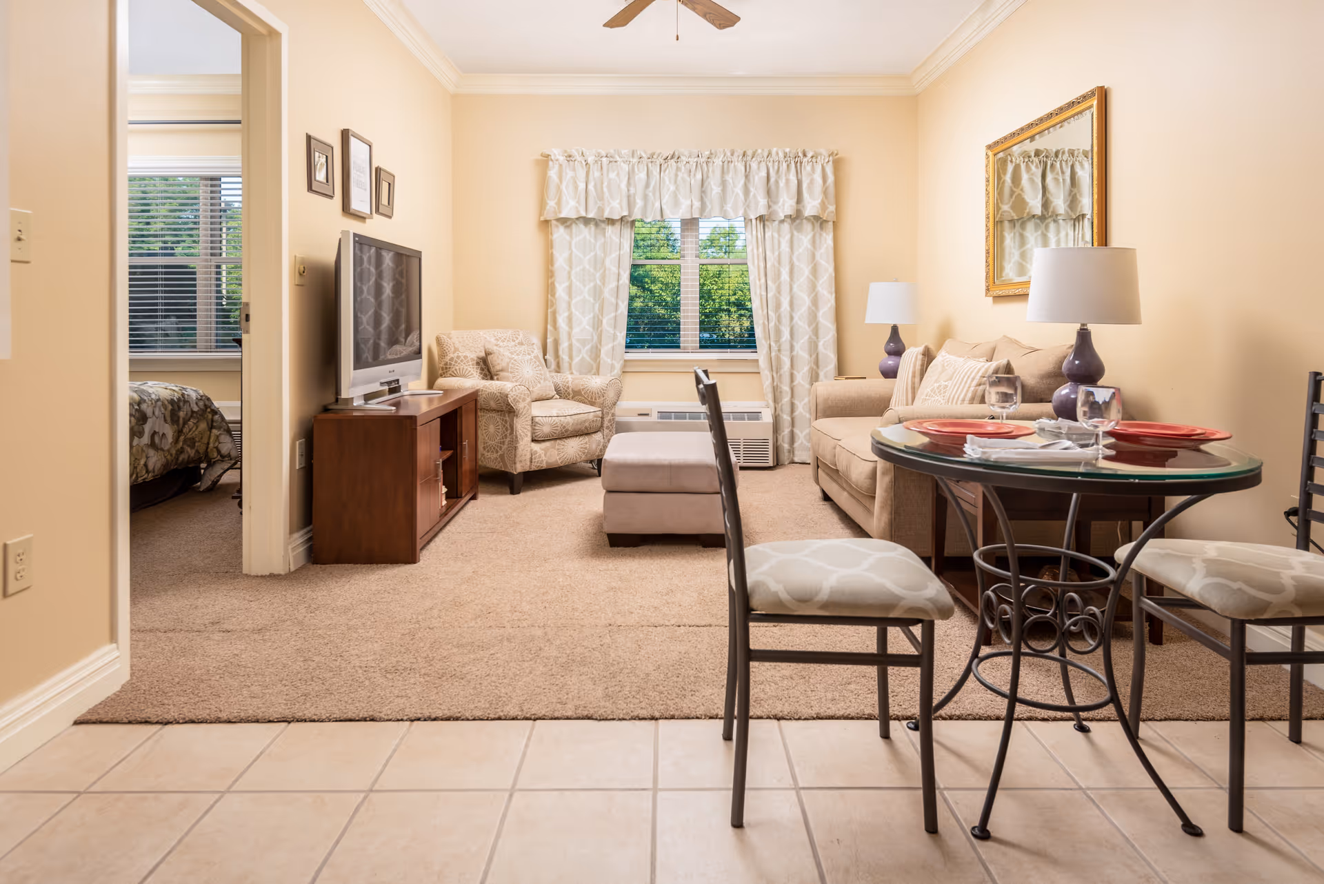 A cozy living area in a senior living facility with beige walls and carpet. The room features a beige sofa with pillows, a patterned armchair, a glass-top dining table set with two chairs, and a TV on a wooden stand. A window with patterned curtains lets in natural light, and a ceiling fan is visible above. An adjacent bedroom with a bed is partially visible through an open doorway.