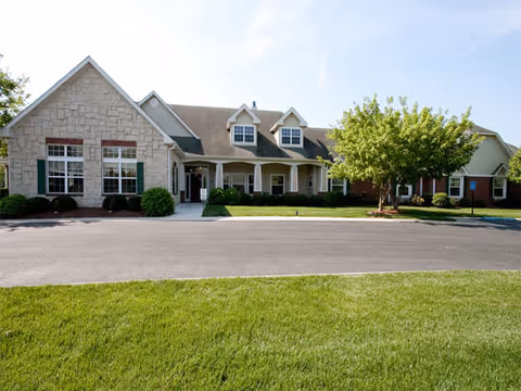 Front exterior view of a single-story senior living facility building with a stone and brick facade, multiple windows, a covered entrance, and a well-maintained lawn and trees in front.
