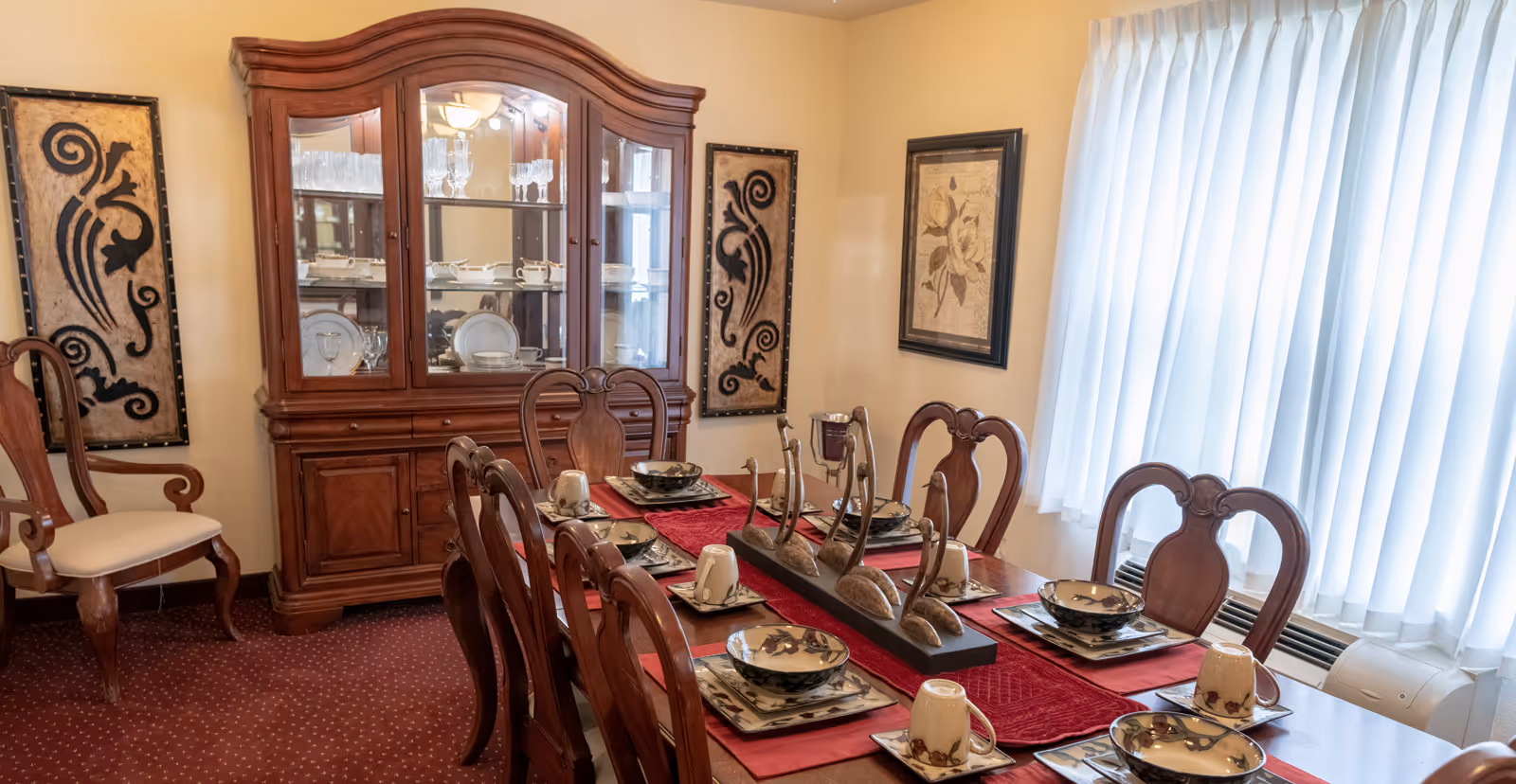Formal dining room with a long wooden table set for a meal, matching chairs, a china cabinet, decorative wall art, and a window with vertical blinds.