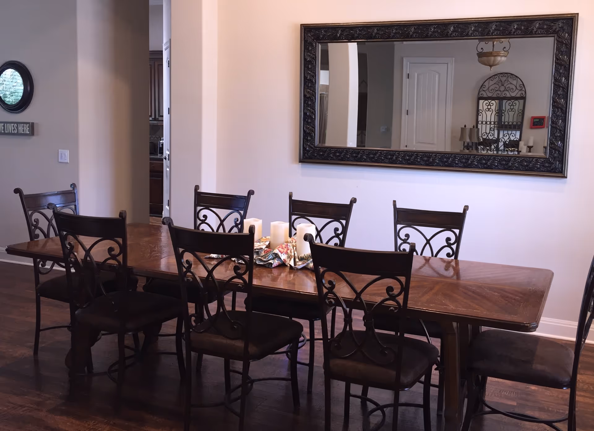 Wood dining table with eight decorative metal-back chairs, candles and a centerpiece, and a large framed mirror on the wall reflecting a hallway.