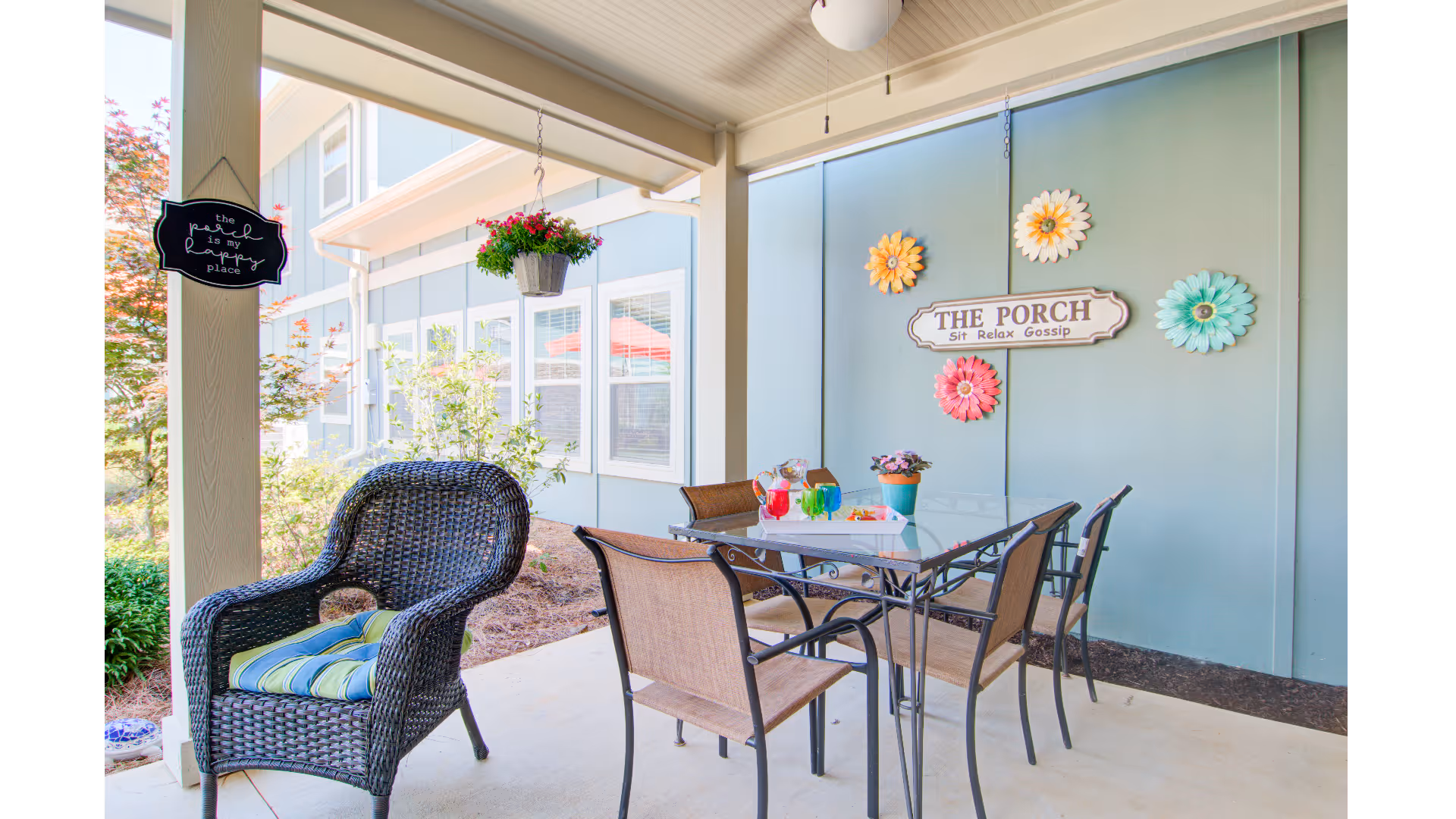 Covered porch with a wicker armchair and glass-top dining table surrounded by chairs, hanging flowers, and a wall sign that reads "THE PORCH."