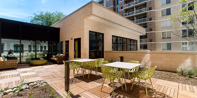 Outdoor patio area with modern green chairs and white tables on a wooden deck, adjacent to a beige building with large windows and a taller apartment building in the background under a blue sky.