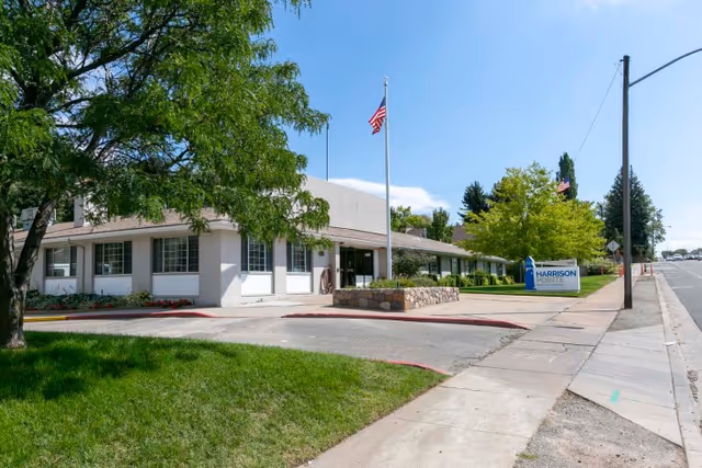 Front exterior of Harrison Pointe Healthcare and Rehabilitation with an American flag, entrance, sidewalk, landscaping and facility sign.