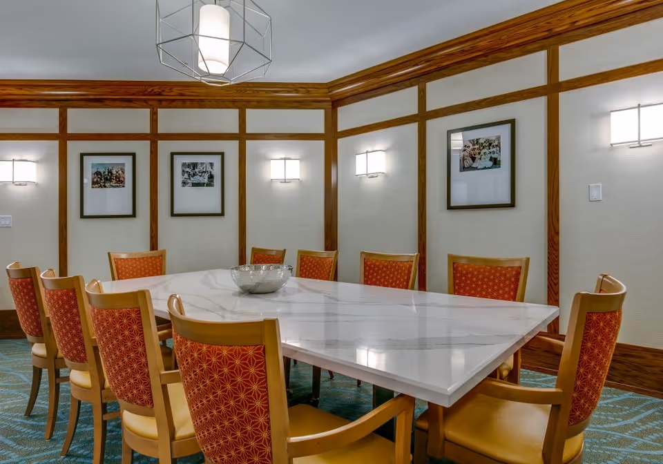 A formal dining room with a large rectangular white marble table surrounded by ten wooden chairs with red patterned upholstery. The walls have wooden paneling with framed pictures and modern wall sconces. A geometric pendant light hangs from the ceiling.