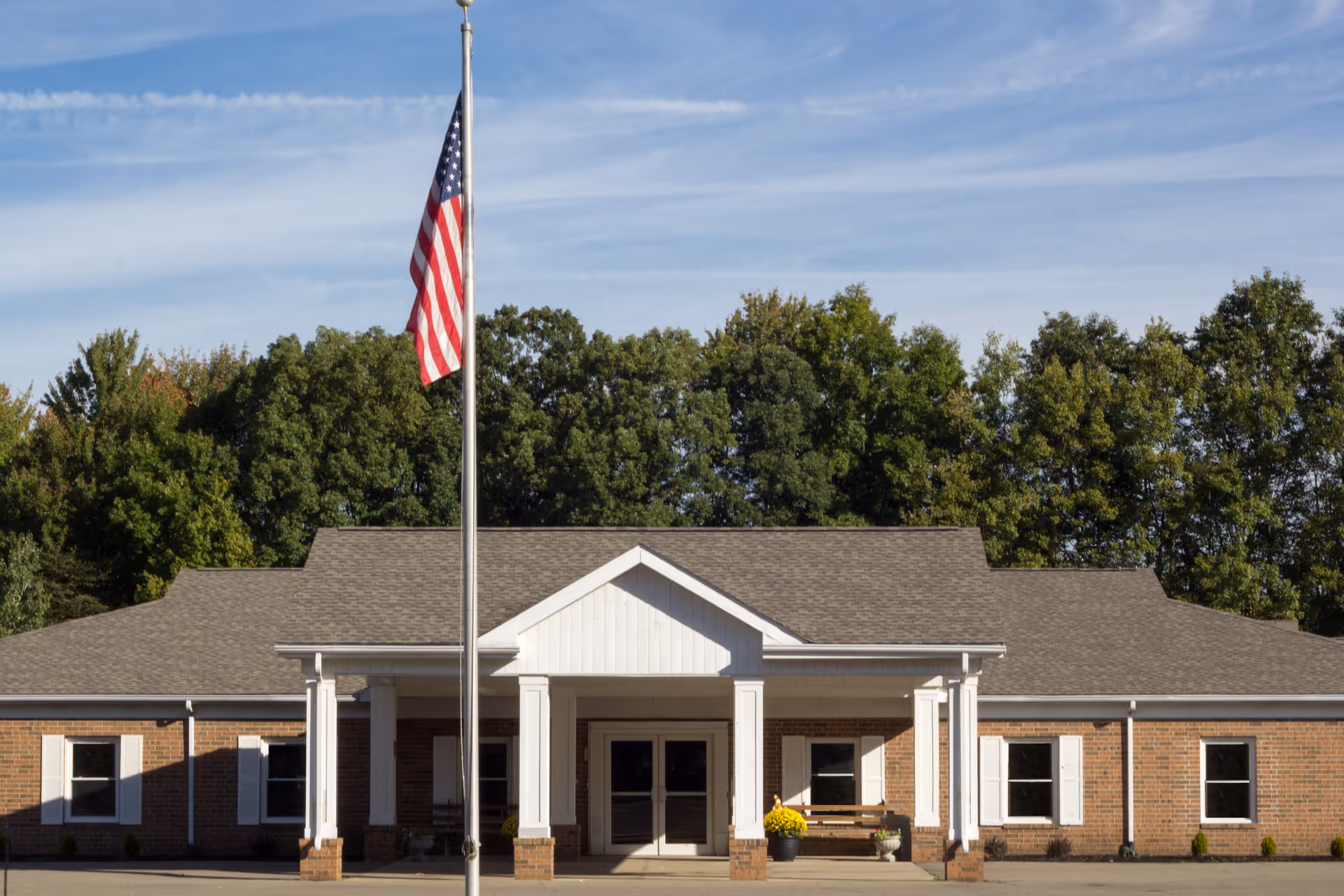 Front exterior view of a single-story brick building with white trim and a covered entrance supported by columns. An American flag on a flagpole is prominently displayed in front of the building, with trees and a blue sky in the background.