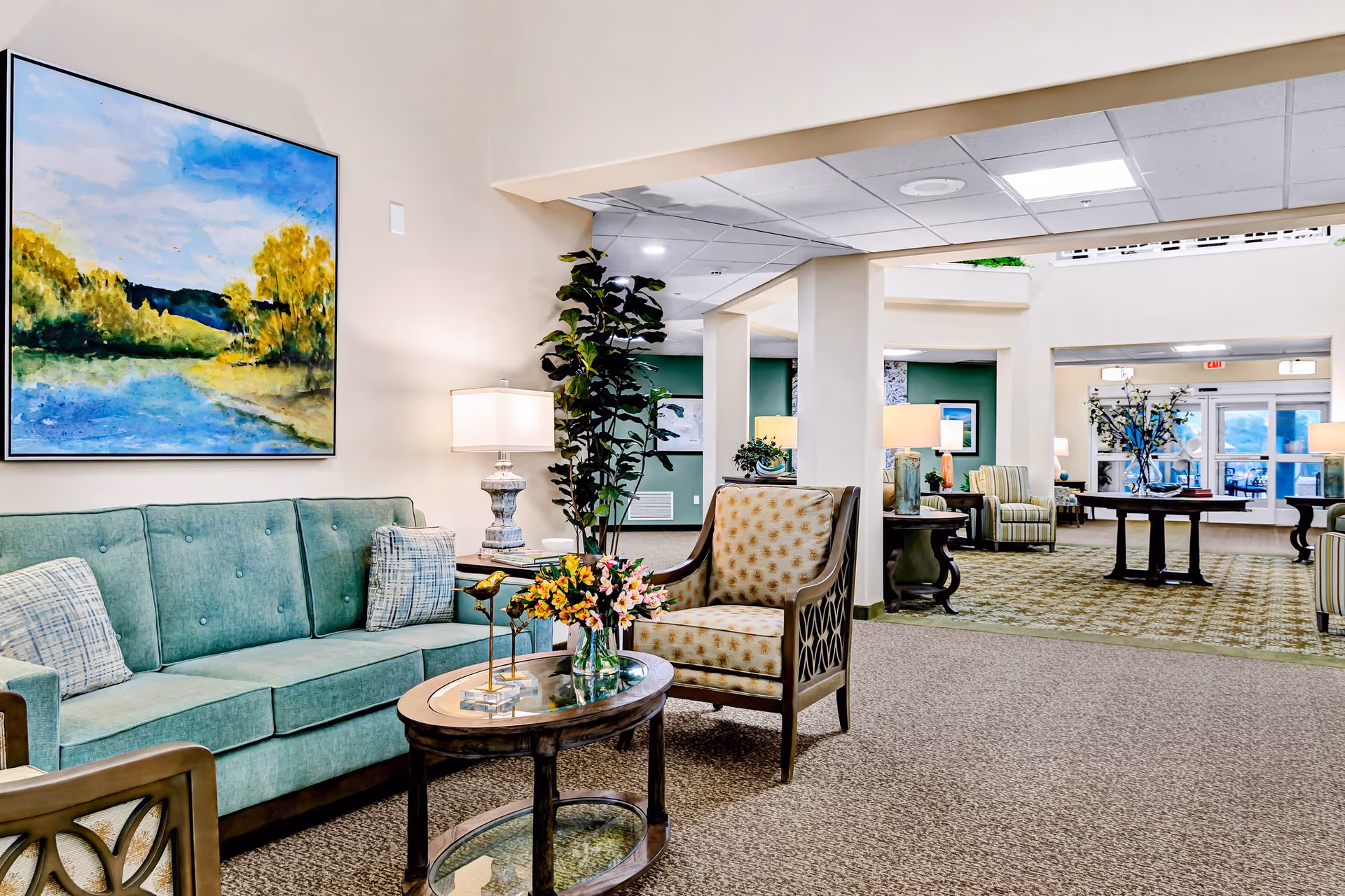 A bright and spacious living room area in a senior living facility featuring a light green sofa with patterned pillows, a wooden armchair with beige patterned upholstery, a glass-top coffee table with a flower vase and decorative bird figurines, a tall potted plant, and a large colorful landscape painting on the wall. The room extends into another seating area with additional chairs, tables, lamps, and large windows letting in natural light.