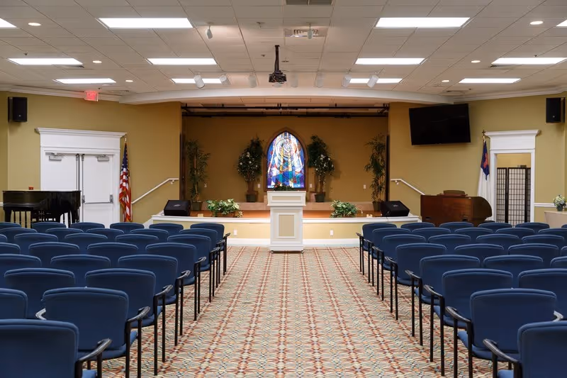 Interior view of a chapel or meeting room with rows of blue chairs facing a small stage with a white podium. Behind the podium is a stained glass window and potted plants. There is a piano on the left side and a TV mounted on the right wall. The room has a patterned carpet and beige walls.