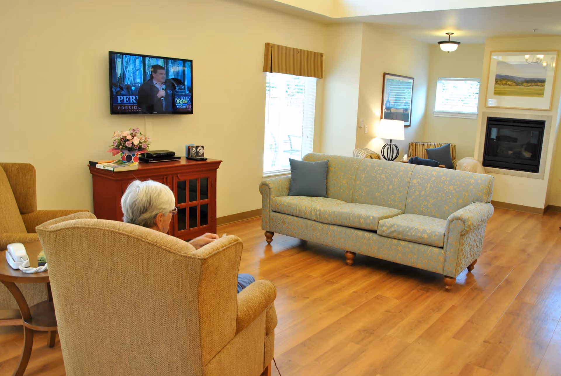 A cozy living room with wooden flooring, featuring a light blue patterned sofa with a blue cushion, a beige armchair occupied by an elderly person with white hair, a small wooden side table with a white telephone, a red cabinet with flowers and books on top, a wall-mounted TV showing a man speaking, and a fireplace with framed artwork above it.