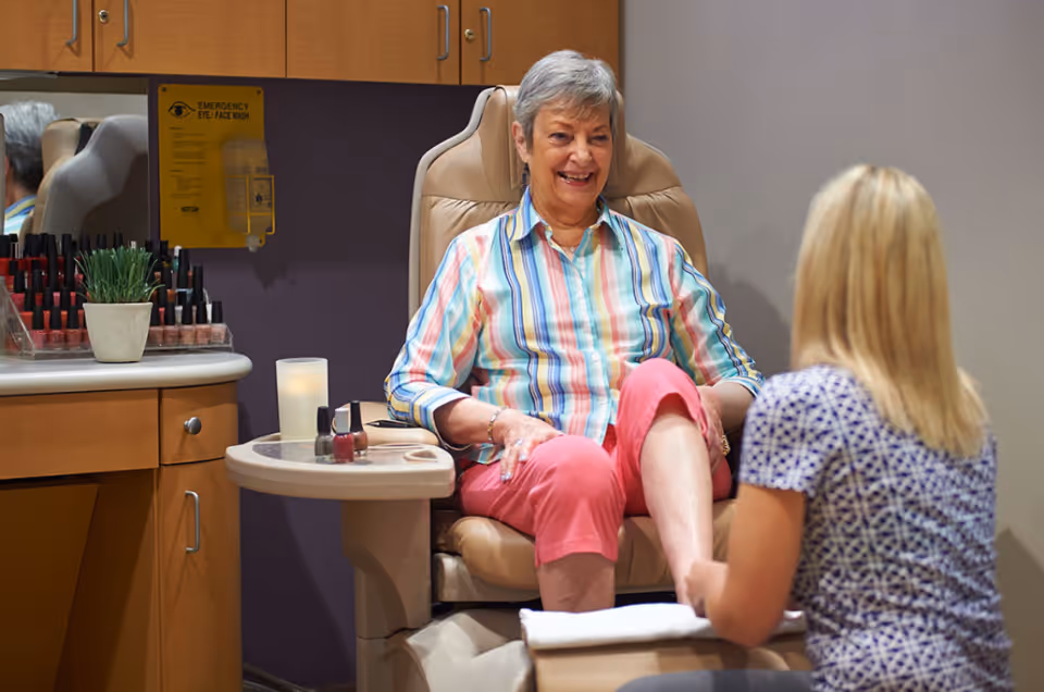 An elderly woman with short gray hair wearing a colorful striped shirt and pink pants is sitting in a beige pedicure chair, smiling while a blonde woman in a patterned shirt is attending to her feet. The setting appears to be a nail salon with nail polish bottles and a small potted plant on a counter nearby.