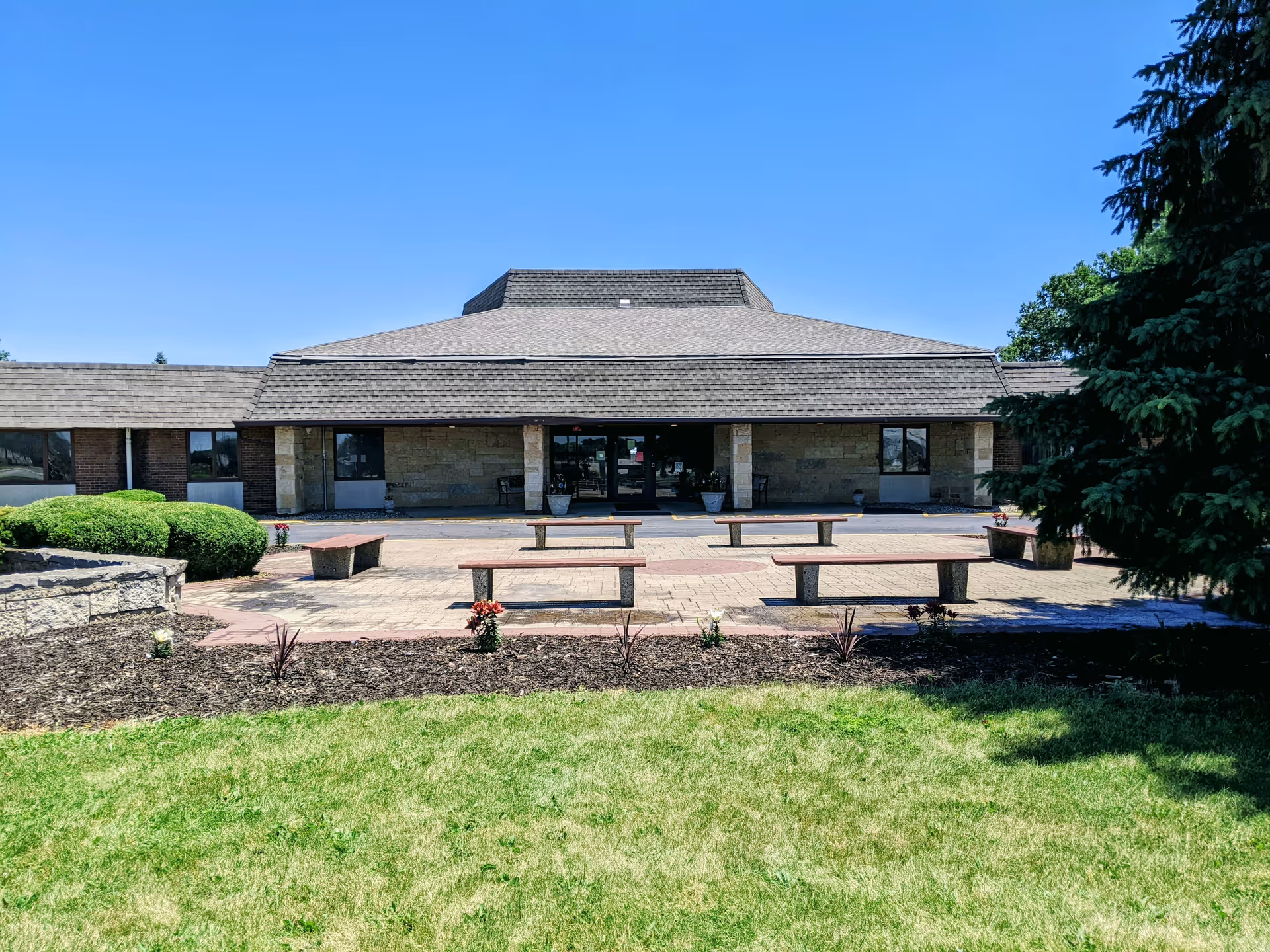 Front exterior view of a single-story building with a large sloped roof, stone and brick facade, and a paved patio area with several wooden benches. There are small flower beds and green bushes in front of the building, and a large tree on the right side. The sky is clear and blue.