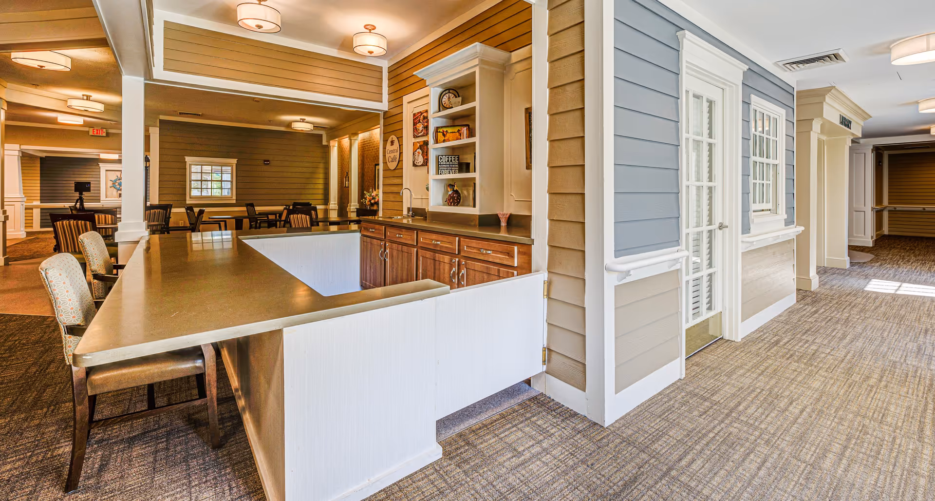Interior view of a senior living facility showing a reception or serving counter with chairs, cabinetry, and a small sink. The area has warm lighting, carpeted floors, and walls with a mix of wood paneling and painted surfaces. In the background, there are tables and chairs arranged for dining or social activities.