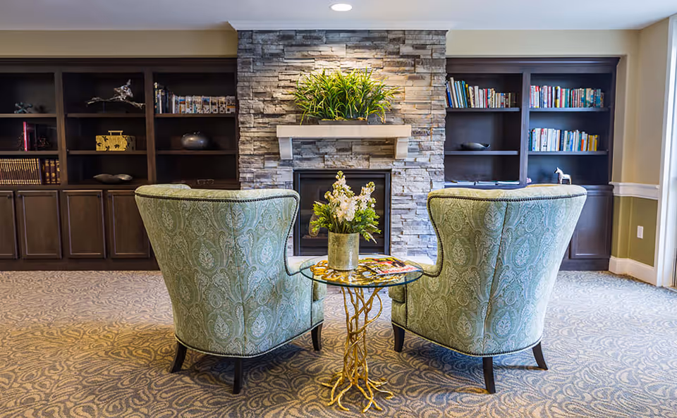 Two patterned armchairs facing a stone fireplace with a green plant on the mantel. Between the chairs is a small glass-top table with a floral arrangement and magazines. Behind the fireplace are built-in dark wood bookshelves filled with books and decorative items. The room has a patterned carpet and neutral-colored walls.