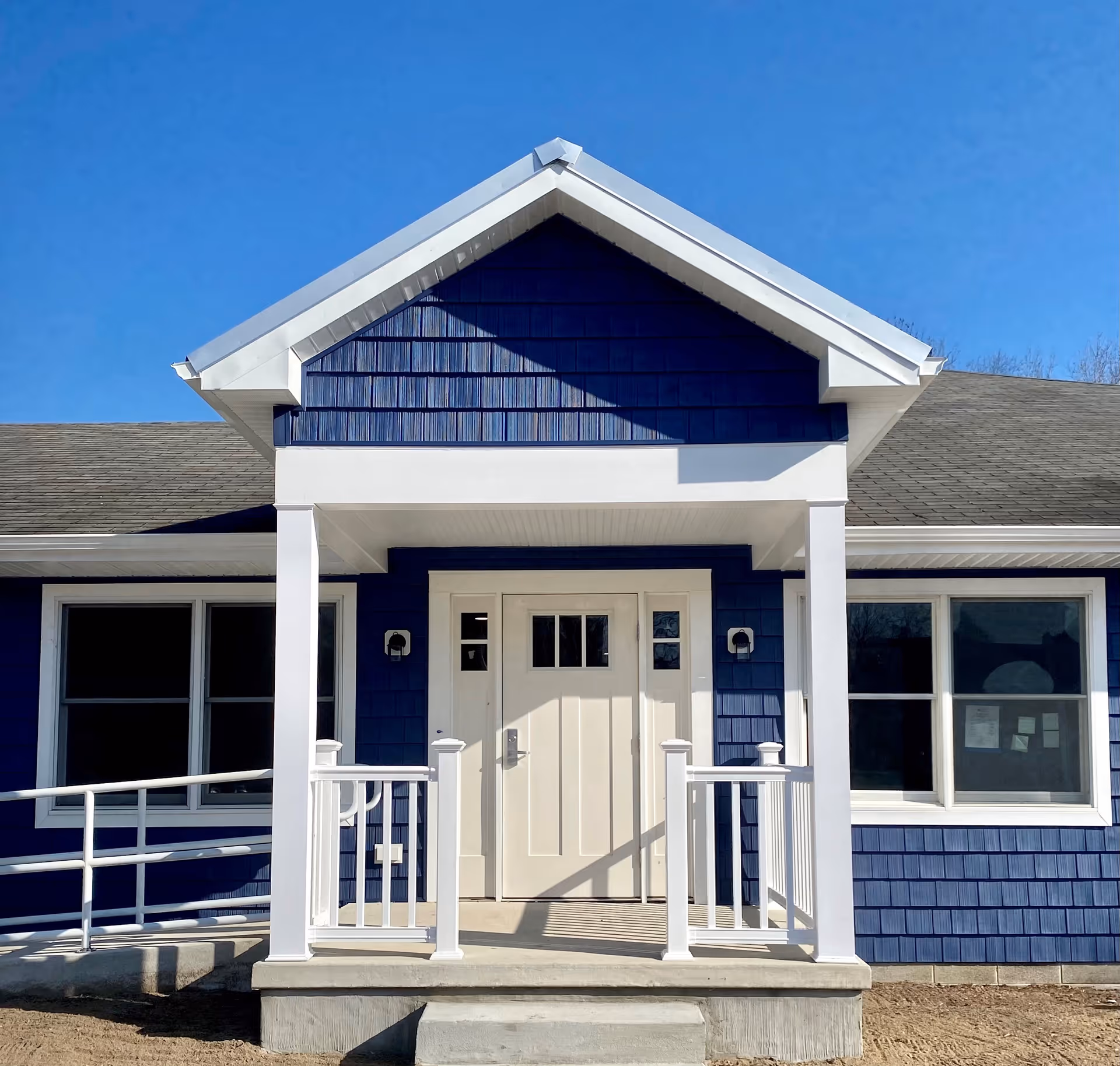 Front entrance of a blue single-story building with a white door, covered porch, columns and an accessibility ramp.