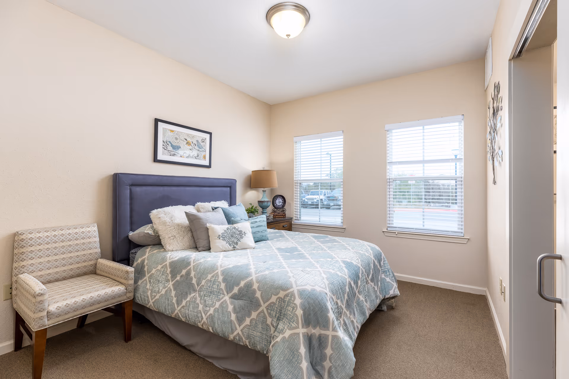 Bright bedroom with a queen bed featuring patterned bedding, a blue headboard, bedside table with a lamp, and a chair by two windows.