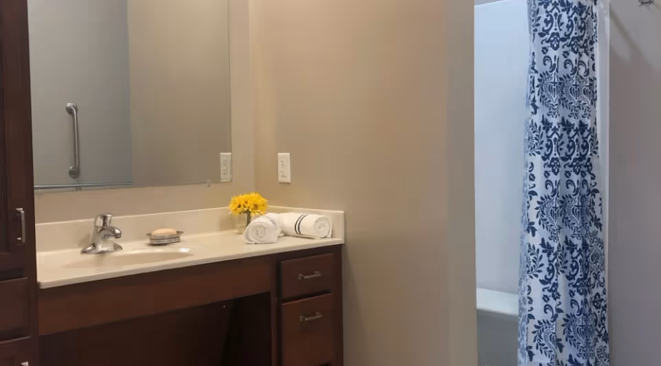 Bathroom interior with a sink and countertop featuring a faucet, soap dish with soap, a small vase with yellow flowers, and two rolled white towels. A large mirror is mounted on the wall above the sink. To the right, a shower area is partially visible with a blue and white patterned shower curtain.