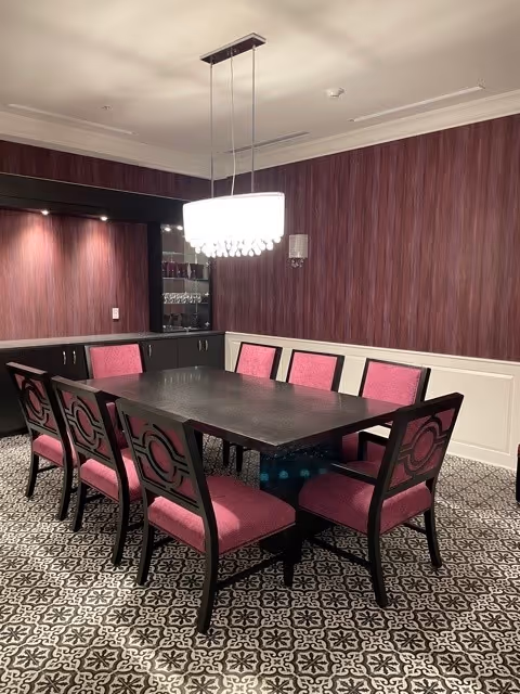 A dining room with a rectangular black table surrounded by eight chairs with pink upholstery and black frames. The room has patterned black and white floor tiles, purple textured wallpaper, white wainscoting, and a modern rectangular chandelier hanging above the table. There is a built-in cabinet with glassware in the background.
