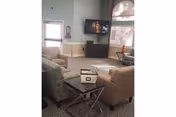 A cozy living room area in a senior living facility with several upholstered chairs arranged around a glass-top side table. A flat-screen TV is mounted on the wall above a cabinet, and there is a window and a glass door letting in natural light.