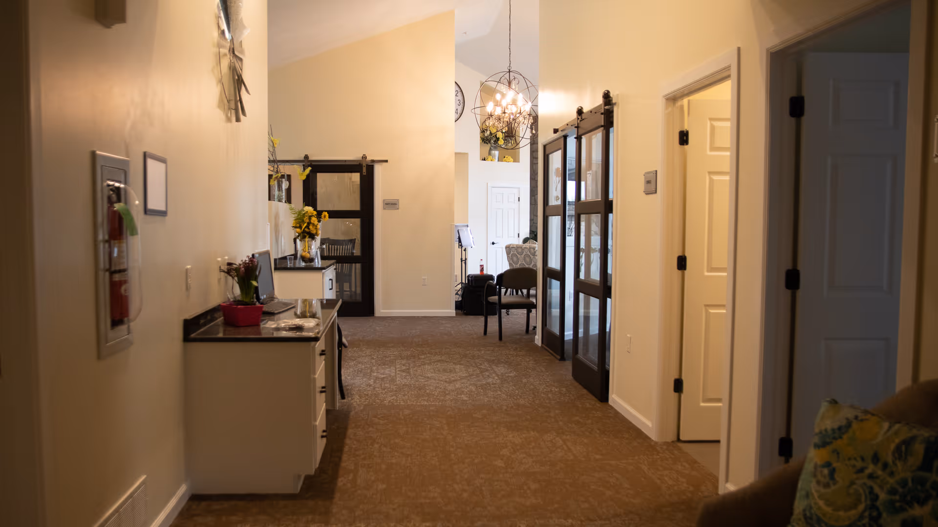 Interior hallway of a senior living facility with beige walls and carpeted floor. On the left side, there is a small counter with drawers, a laptop, and decorative flowers. On the right side, there are open doorways leading to other rooms. At the end of the hallway, there are dark wooden sliding doors and a chandelier hanging from the ceiling. A chair and part of a patterned armchair are visible in the background.