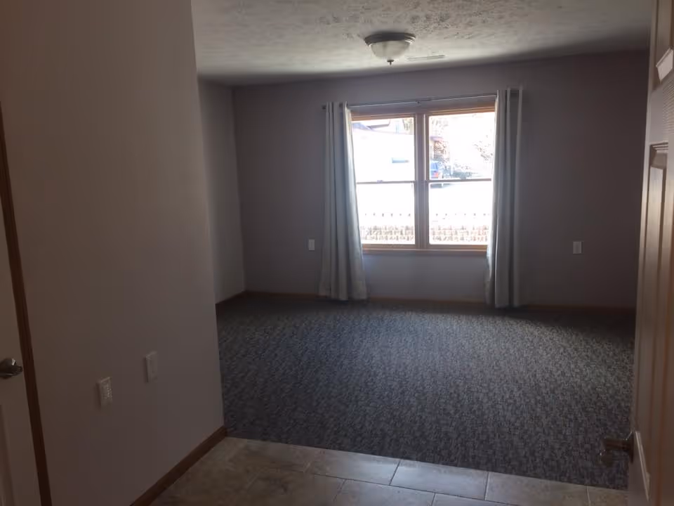 Empty carpeted room with a window and light curtains, a tile entryway, and a ceiling light fixture.
