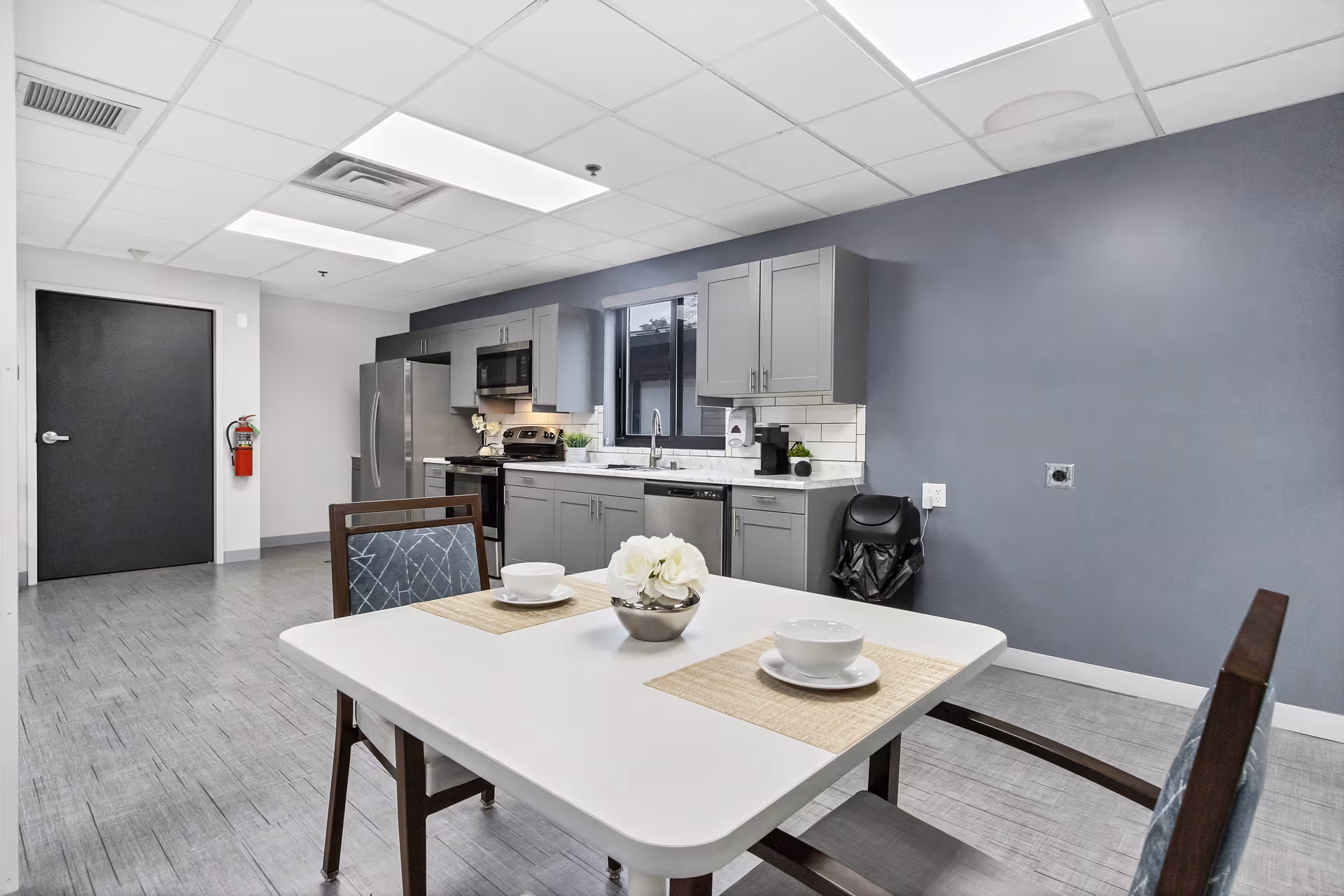 A modern kitchen and dining area with gray cabinets, stainless steel appliances including a refrigerator, stove, microwave, and dishwasher. A white table with two place settings and a small flower arrangement is in the foreground. The walls are painted gray and white, with a window above the sink. The floor is covered with gray tiles, and there is a black door and a fire extinguisher on the left side.