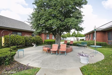 Outdoor courtyard area with a large tree in the center surrounded by a circular brick planter. Several cushioned chairs and metal tables are arranged around the tree on a concrete patio. The courtyard is bordered by red brick buildings and green shrubs, with a white fence and a lamppost visible in the background under a partly cloudy sky.