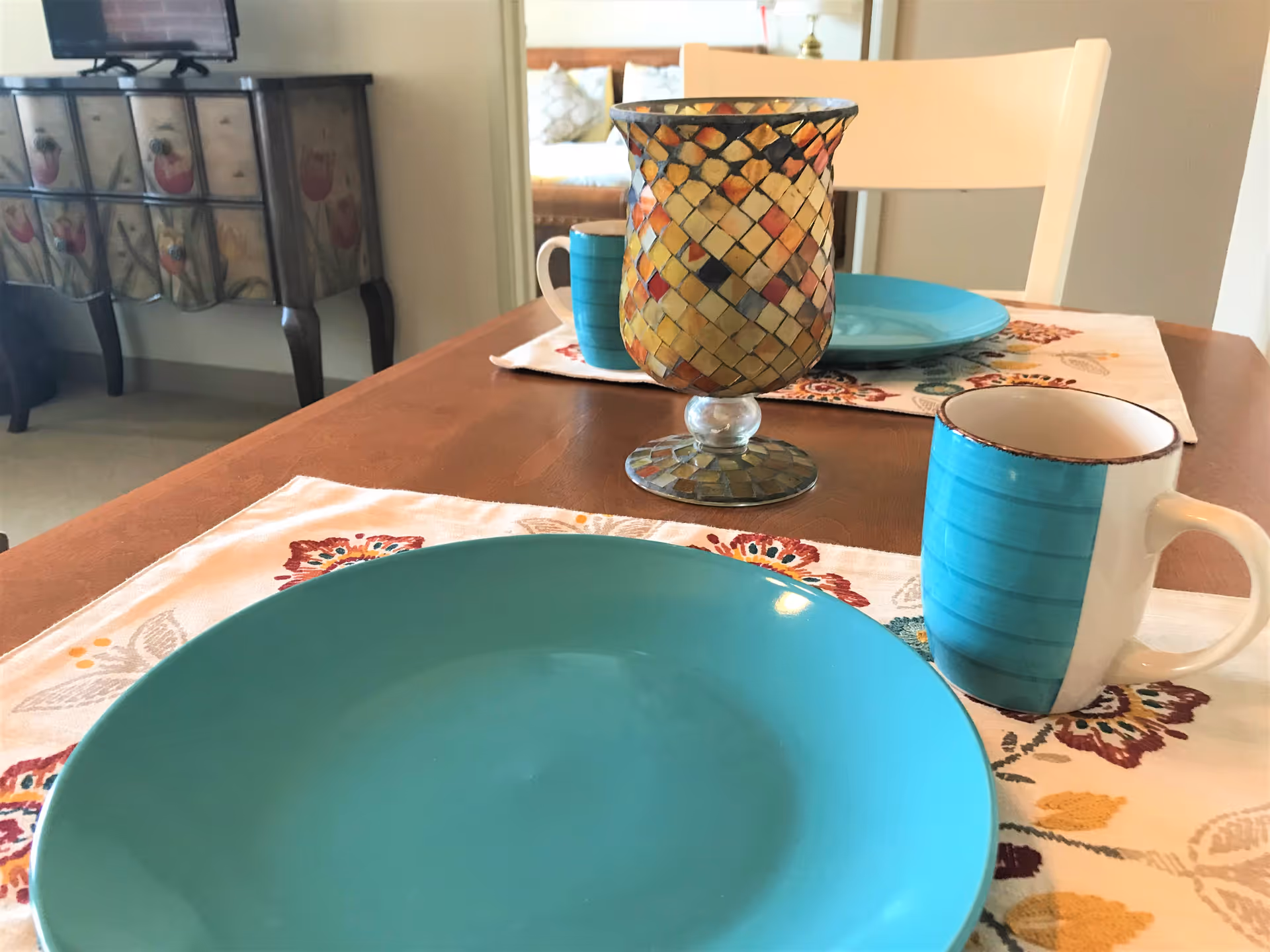 Dining table in a senior living apartment set with turquoise plates and mugs, a mosaic glass centerpiece, and floral placemats.