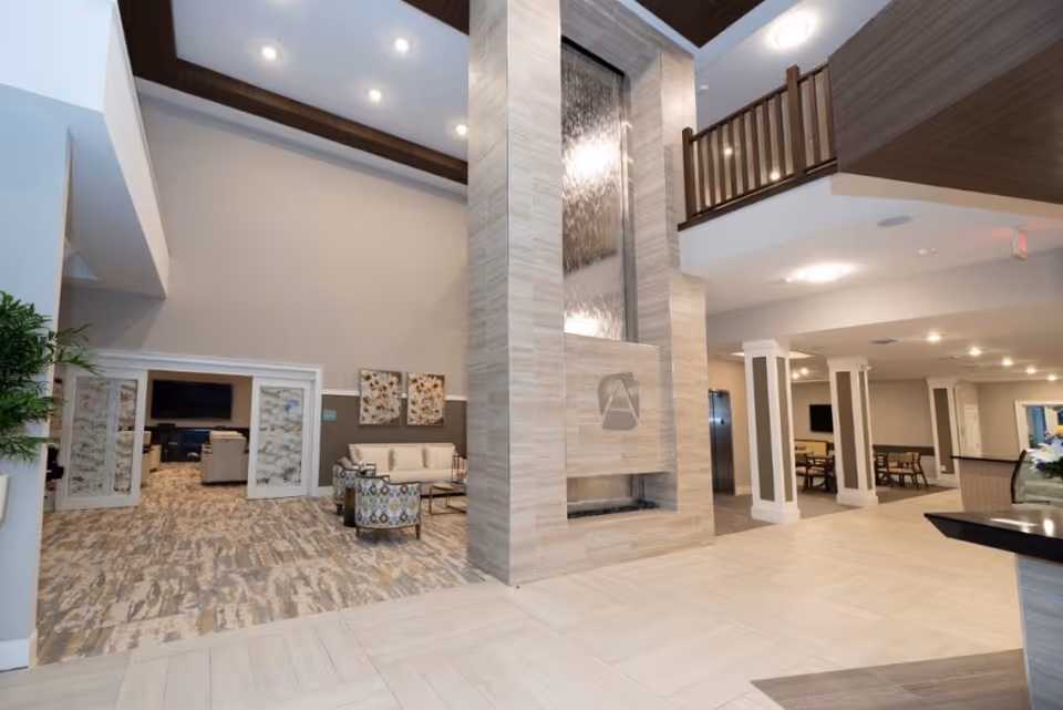 Spacious and modern interior of a senior living facility featuring a tall water feature with the Aspire At West End logo, seating area with chairs and a sofa, decorative wall art, and a view into an office space through double doors. The area has high ceilings with recessed lighting and a balcony railing above.
