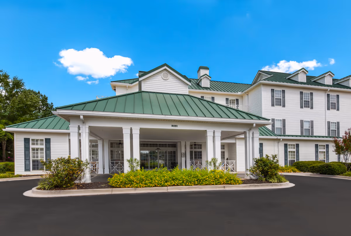 Front exterior view of Arbor Terrace Knoxville, a large white building with green metal roofing, multiple windows with shutters, and a covered entrance with white pillars surrounded by landscaped bushes and flowers under a blue sky with a few clouds.