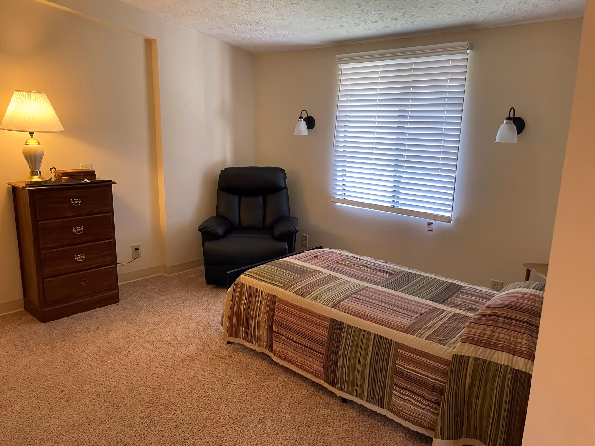 A bedroom with a single bed covered in a striped quilt, a black recliner chair, a wooden chest of drawers with a lamp on top, and a window with closed blinds. The walls are light-colored and the floor is carpeted.