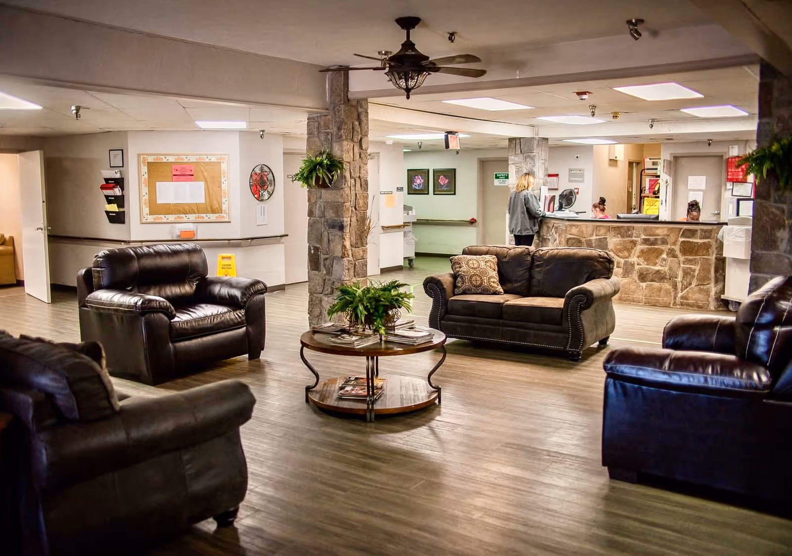 A cozy nursing home common area with several dark leather armchairs and a sofa arranged around a round wooden coffee table with plants and magazines. The room has wood flooring, stone pillars, and a reception desk in the background where a staff member is assisting a visitor.