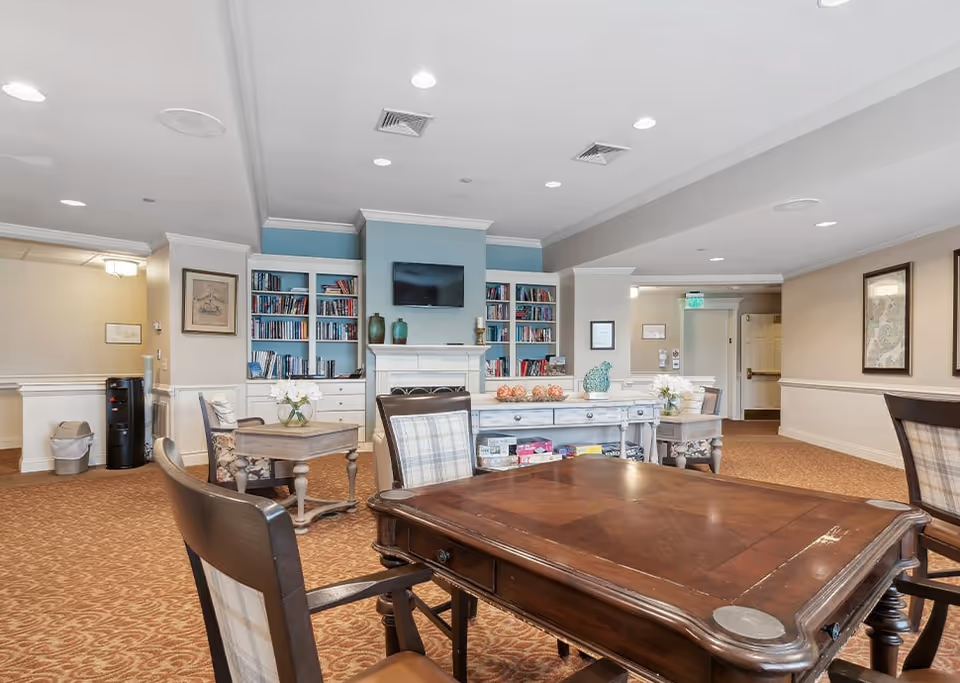 A cozy senior living common area with wooden tables and chairs, a fireplace with a mounted TV above it, built-in bookshelves filled with books, and decorative items on a sideboard. The room has beige walls, carpeted floors, and recessed ceiling lights.