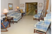 A seating area along a facility corridor with plaid armchairs and sofas, side tables with lamps, and framed pictures on the walls.