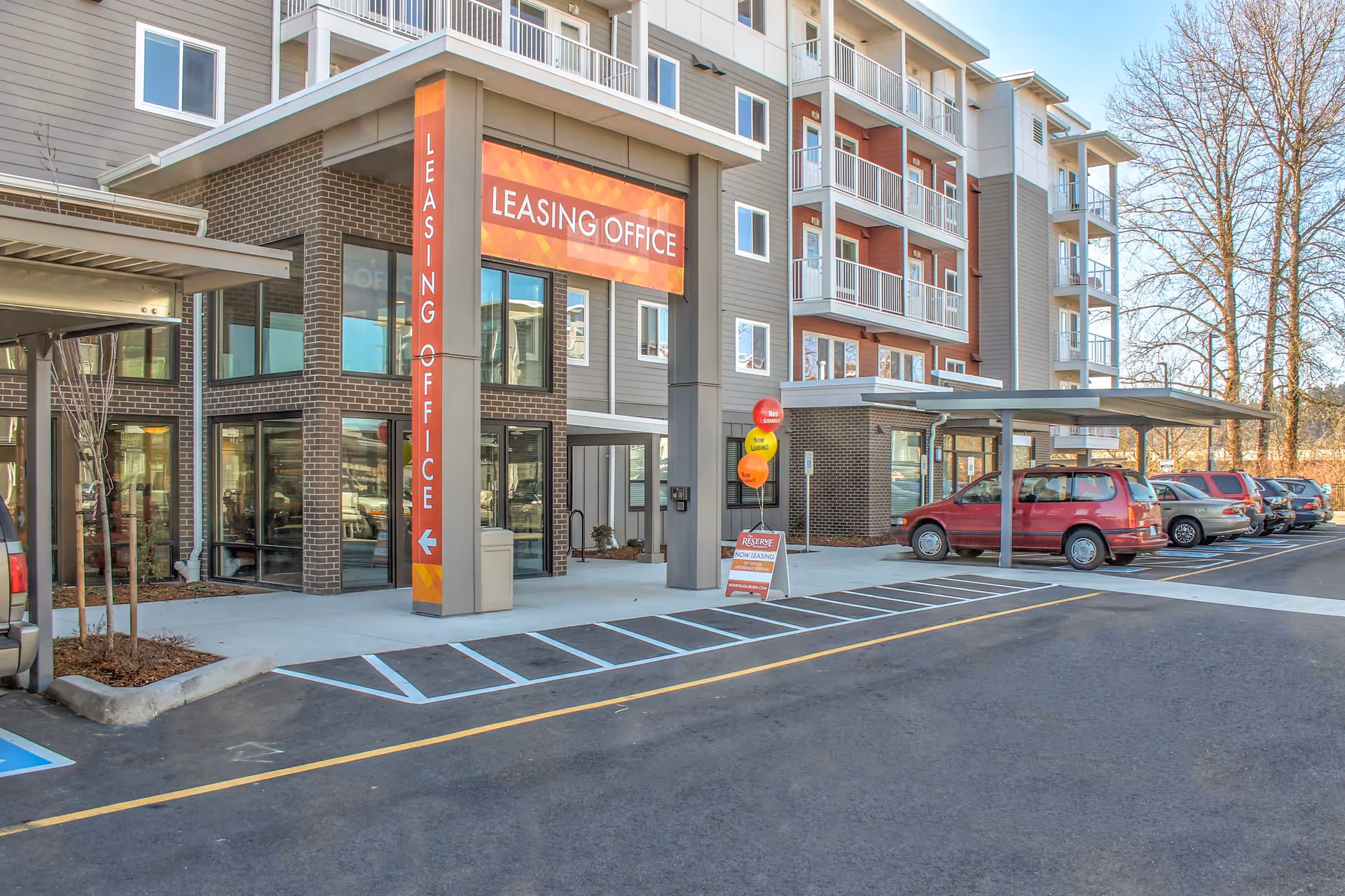 Exterior view of a modern multi-story apartment building showing the leasing office entrance with a sign, balloons, and parked cars.