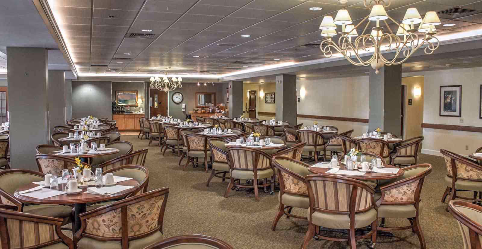 A spacious dining room with multiple round tables, each set with white cups, glasses, napkins, and small flower arrangements. The room features upholstered chairs with floral patterns, chandeliers hanging from the ceiling, and a buffet area with a clock on the wall in the background.