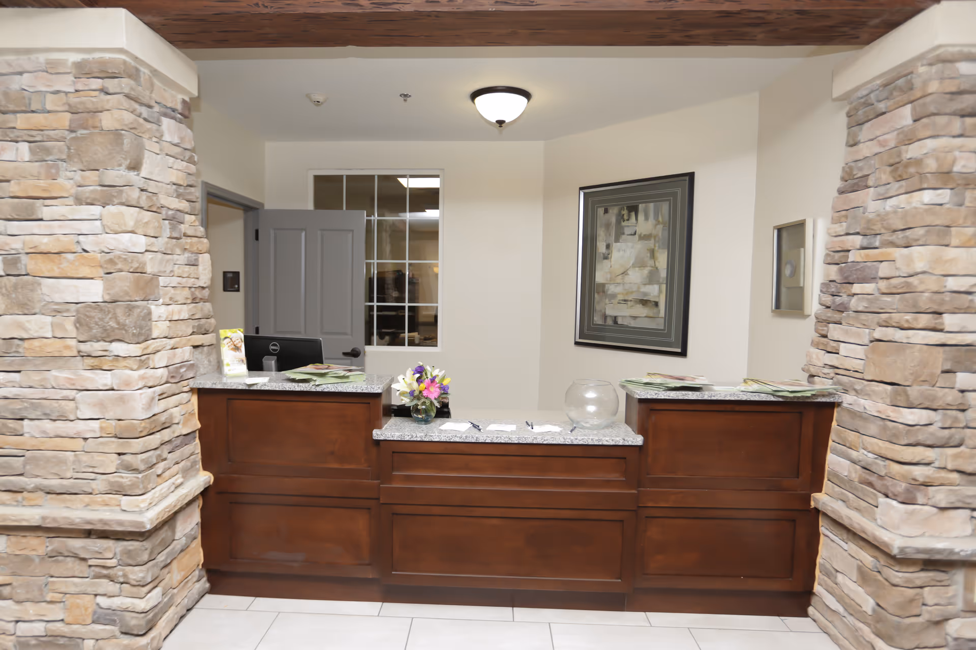 Reception desk area with a dark wooden counter and granite top, flanked by two stone pillars. There is a small vase with flowers, some papers, and a glass bowl on the counter. Behind the desk is a closed gray door and a window with multiple panes. A framed abstract painting hangs on the wall.