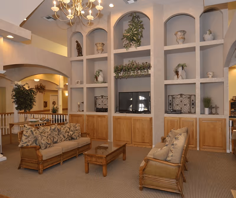 A cozy living room area in a senior living facility featuring two wooden sofas with floral cushions, a wooden coffee table, and built-in shelves with decorative plants and ornaments. The room has beige walls, carpeted flooring, and a chandelier hanging from the ceiling.