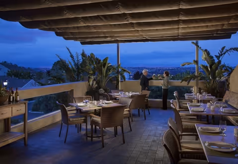 Outdoor dining area at dusk with several tables and chairs set for a meal. Two people are standing and conversing near the railing, with a scenic view of trees and city lights in the background. Large plants and an overhead canopy are also visible.