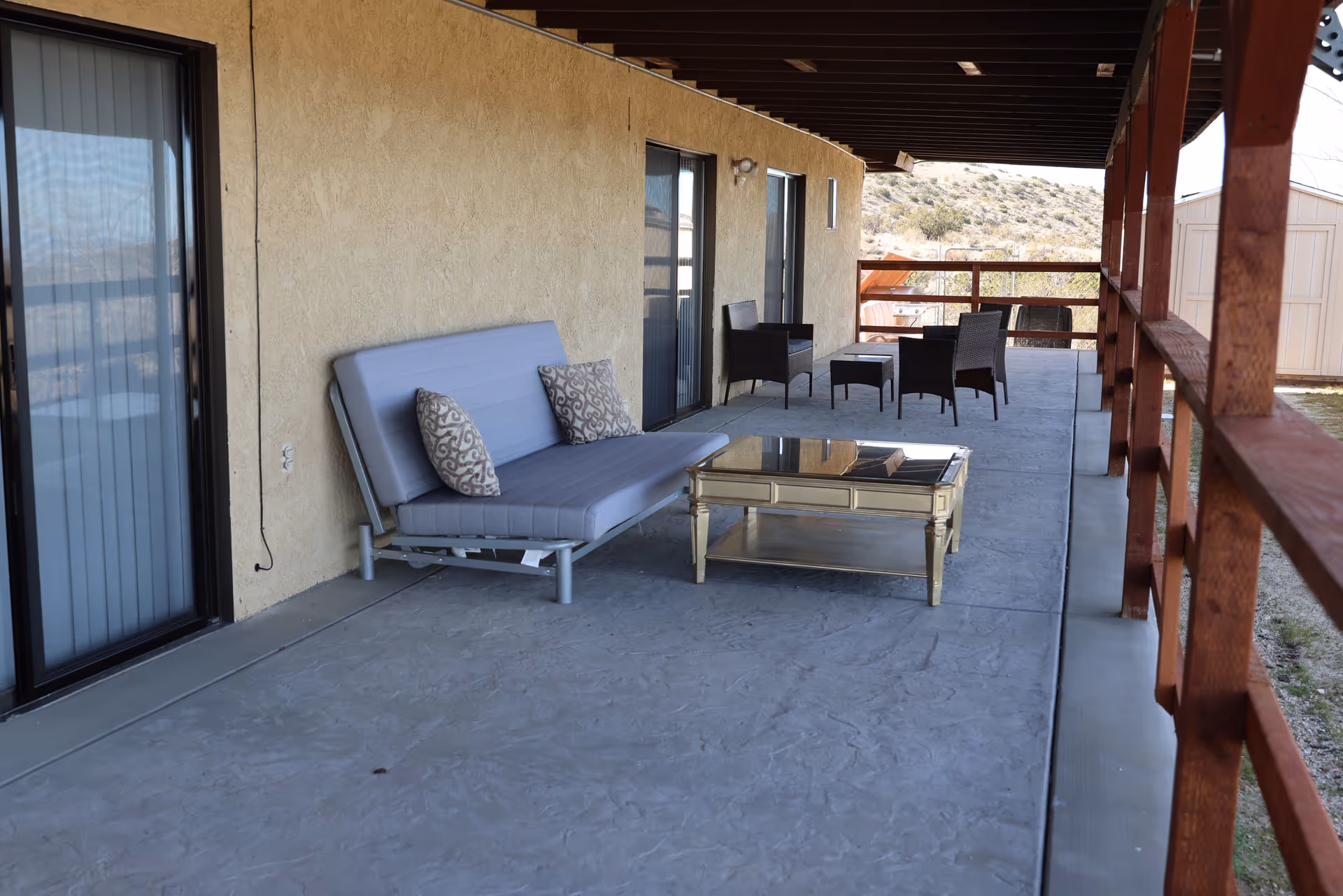 Covered outdoor patio area with a gray cushioned bench with two patterned pillows, a glass-top coffee table, and a set of dark wicker chairs with a small table. The patio has a concrete floor and wooden railing, with a view of a hilly landscape in the background.