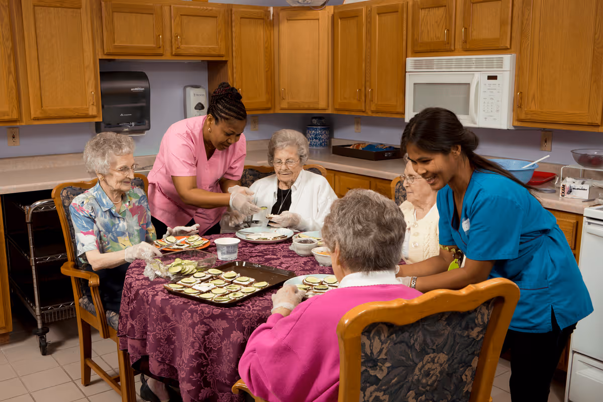 Two caregivers assist four elderly women seated around a table in a kitchen, preparing snacks with sliced cucumbers and other ingredients on a tray. The kitchen has wooden cabinets, a microwave, and a paper towel dispenser on the wall.