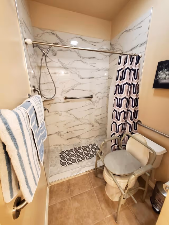 Bathroom with a walk-in shower with marble-pattern walls, grab bars, shower curtain, and a raised toilet seat with support frame.
