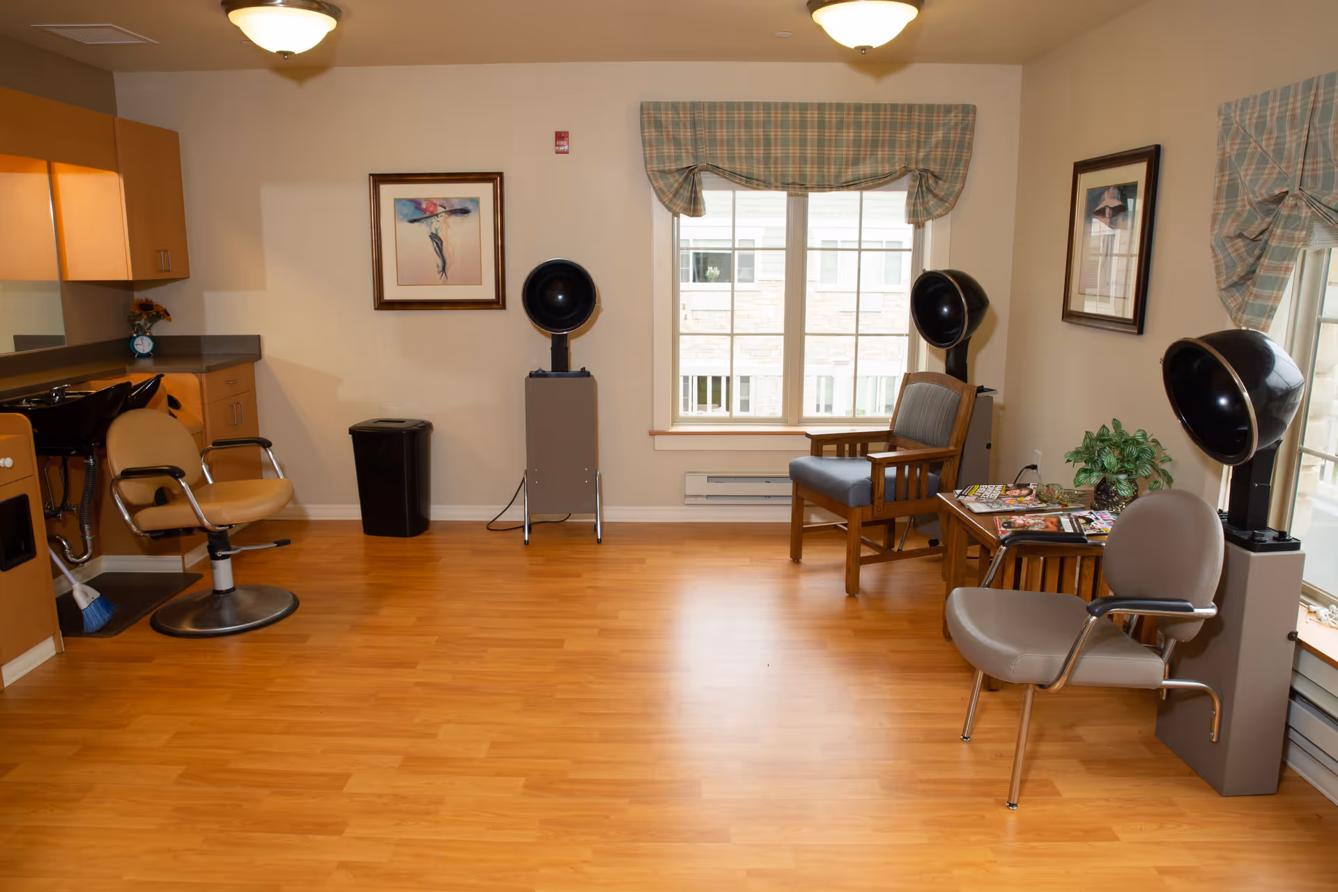 Interior of a hair salon area in a senior living facility with wooden flooring, two salon chairs, hair drying stations, a small table with magazines, a wooden armchair, framed artwork on the walls, and a large window with plaid valances.