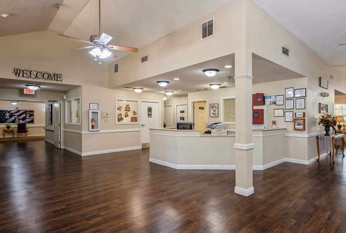 Interior view of a senior living facility reception area with a front desk, ceiling fans, framed certificates on the wall, and a 'WELCOME' sign above a hallway entrance. The space has wood flooring, beige walls, and overhead lighting.
