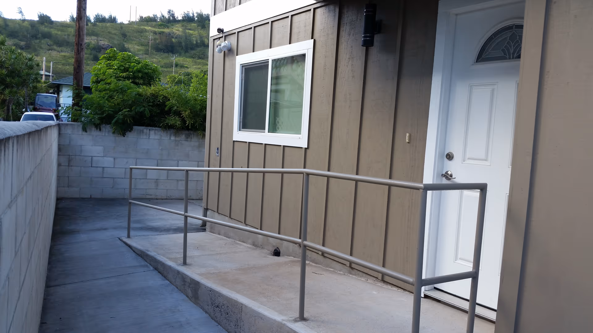 Concrete wheelchair ramp with metal handrails leading to a white door on the exterior of a beige building with a cinder block wall and greenery in the background.