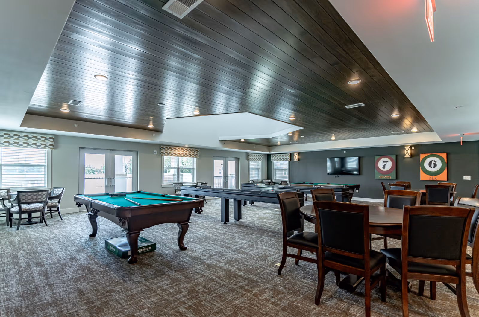 A spacious game room with a pool table, shuffleboard table, multiple seating areas with tables and chairs, a wall-mounted TV, and decorative artwork featuring billiard balls numbered six and seven. The room has large windows and glass doors allowing natural light, a dark wood-paneled ceiling with recessed lighting, and carpeted flooring.