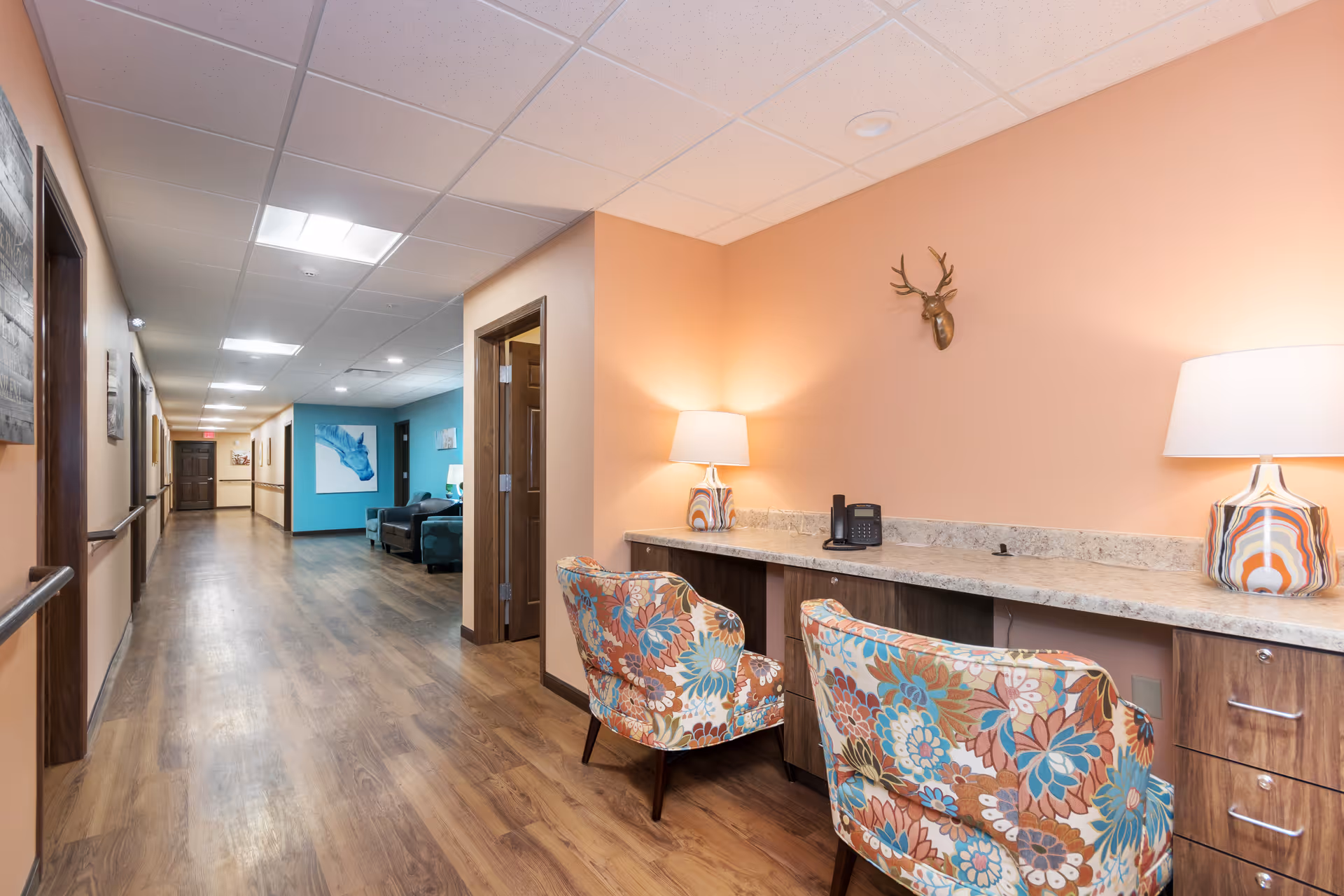 Interior hallway of a memory care facility with a built-in desk area, two floral chairs, table lamps, and a long wood-floored corridor leading to a seating nook.