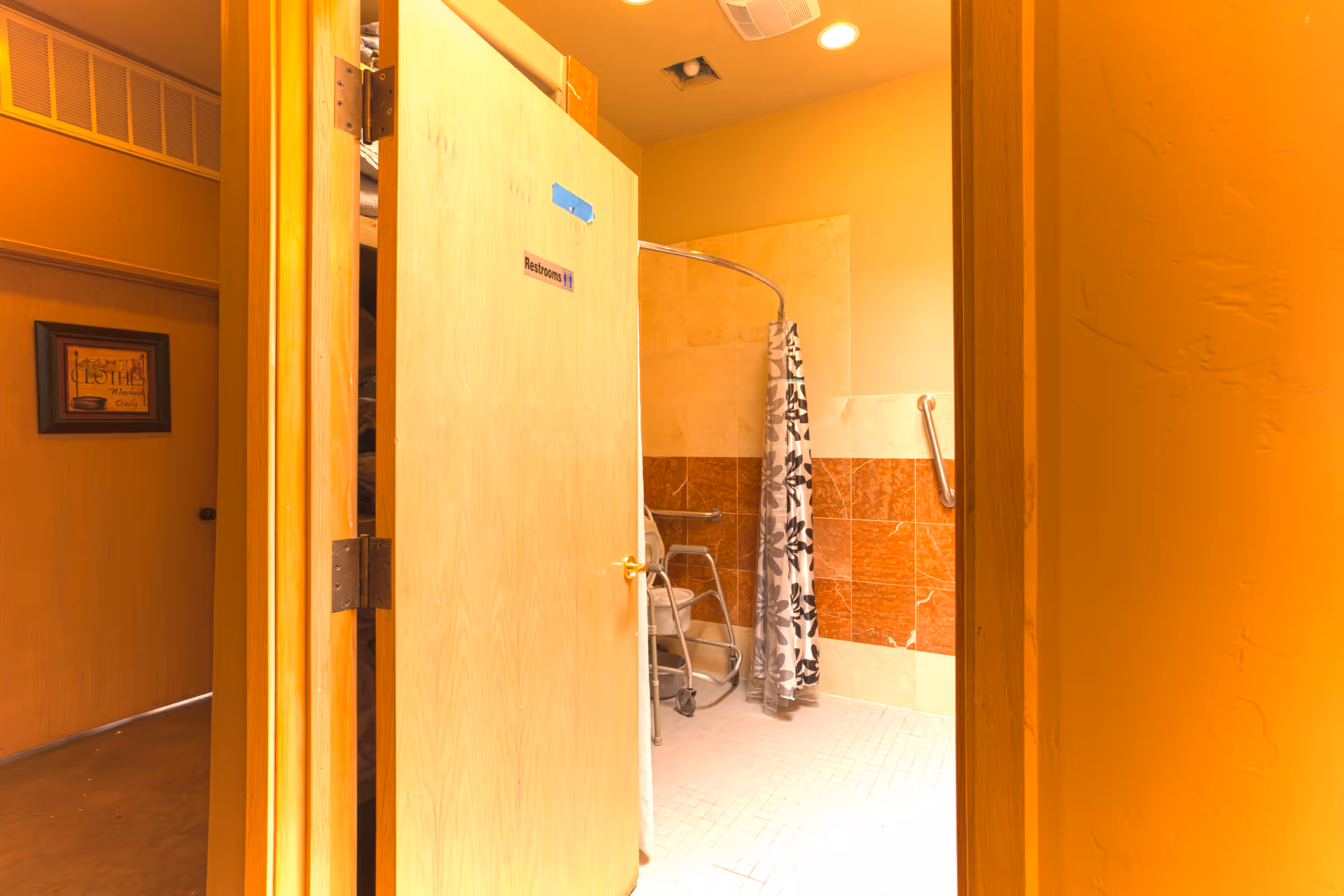 View through an open door into a restroom with a walk-in shower area that has a floral shower curtain and a walker inside. The restroom walls are tiled with beige and reddish-brown tiles, and there are grab bars installed for accessibility. The door has a small sign that reads 'Restrooms'.