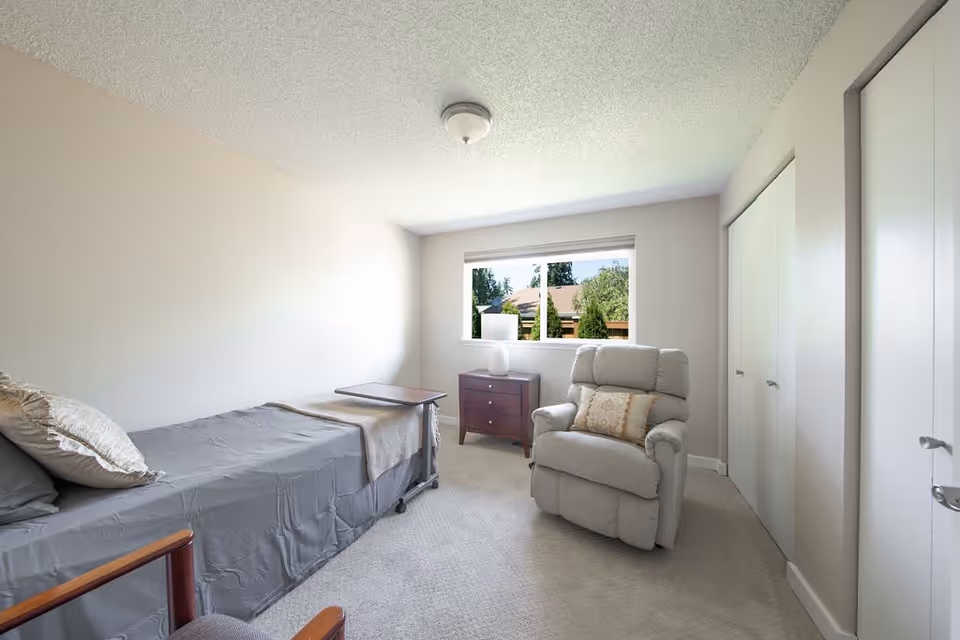 A small, simple bedroom with a single bed covered in gray bedding and a beige throw blanket. There is a wooden side table with a lamp next to the window, and a comfortable beige recliner chair with a decorative pillow. The room has light-colored walls, a window showing greenery outside, and a closet with white sliding doors.