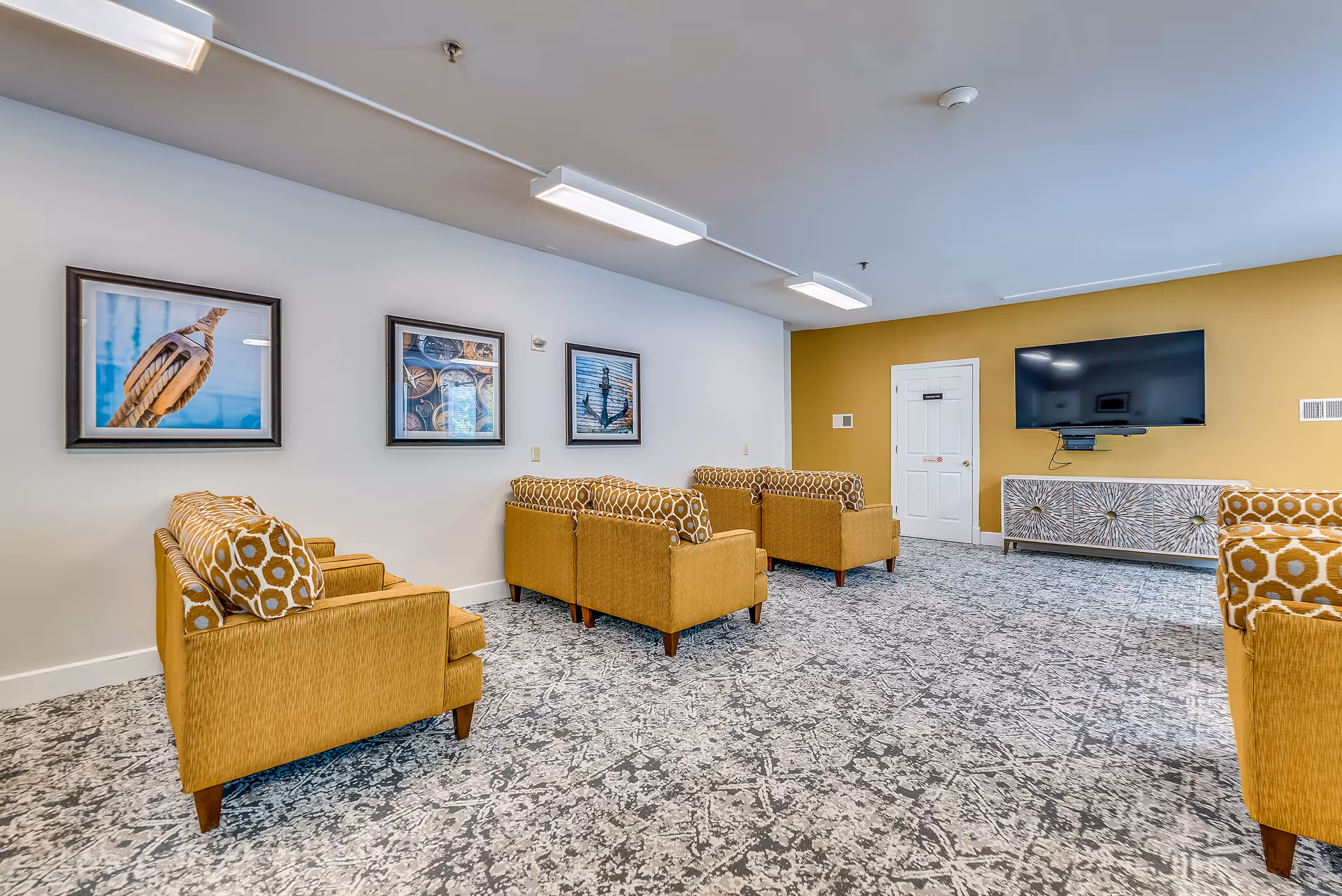 A lounge area in Guilford Retirement Residence featuring mustard yellow armchairs with patterned cushions arranged on a gray and white patterned carpet. The walls are painted white and mustard yellow, decorated with three framed nautical-themed pictures. A flat-screen TV is mounted on the mustard yellow wall above a decorative console cabinet. The room is lit by rectangular ceiling lights.