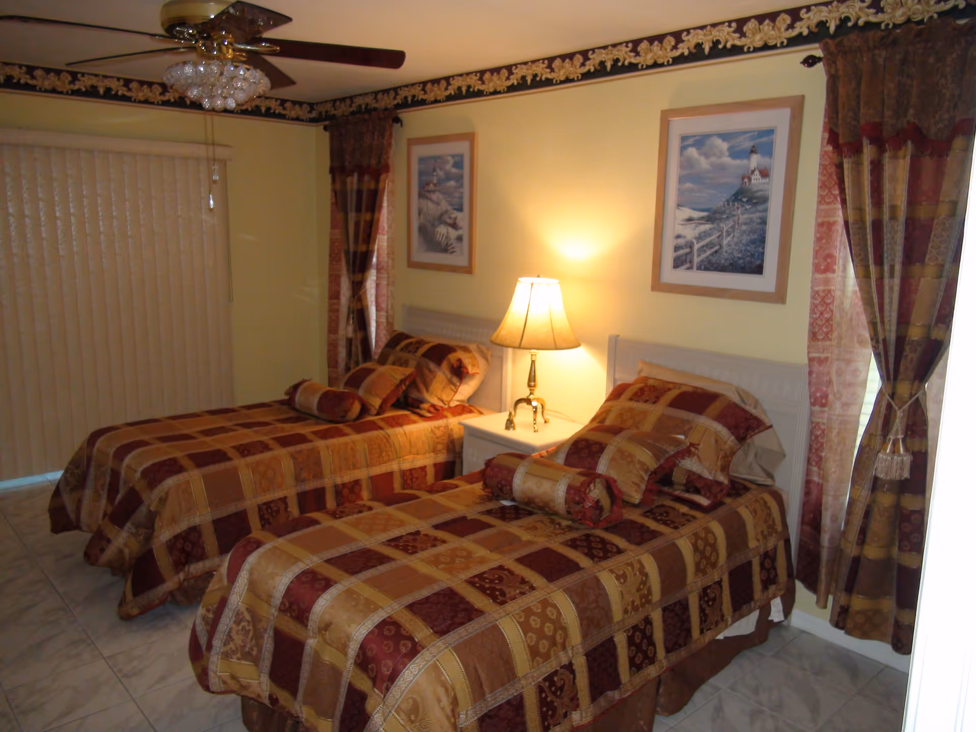 A bedroom with two twin beds covered in matching patterned bedspreads in shades of brown, gold, and burgundy. Each bed has multiple pillows and a bolster pillow. Between the beds is a white nightstand with a lamp emitting a warm light. The walls are painted yellow and decorated with two framed pictures. The room has tiled flooring, a ceiling fan with a light fixture, and windows with patterned curtains and vertical blinds.